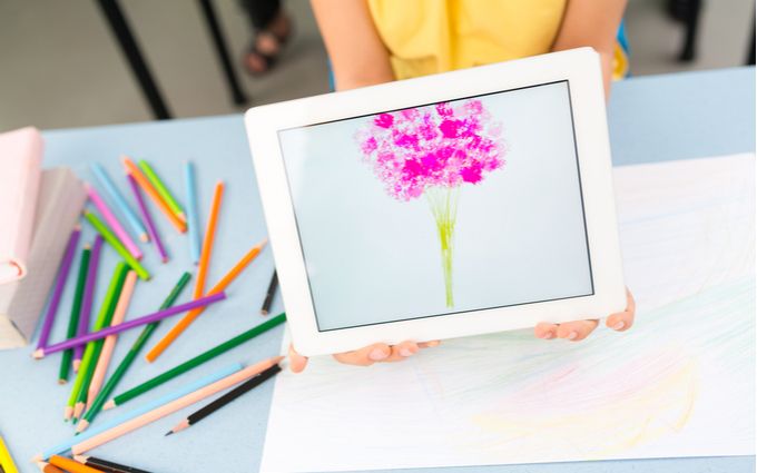 花の絵が描かれたタブレットを持つ人間の横でカラフルな色鉛筆が散らばっている画像